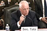 A man wearing business attire sits at a table and speaks into a microphone; in front of him is a place card that reads, "Sec. Tata."
