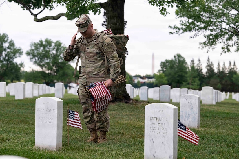 A man in a camouflage military uniform stands in a cemetery and salutes toward a gravesite, while holding small American flags.