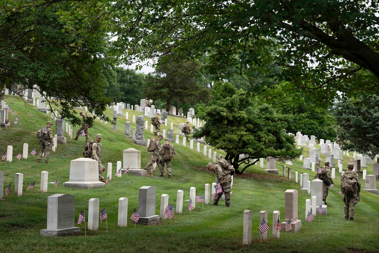 Nearly a dozen people in camouflage military uniforms walk through a cemetery, placing small American flags at each gravesite.