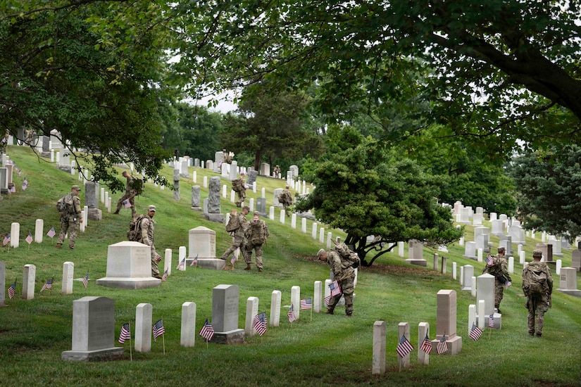 Nearly a dozen people in camouflage military uniforms walk through a cemetery, placing small American flags at each gravesite.