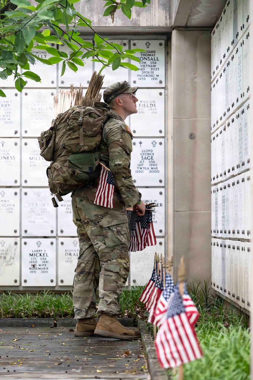 A man in a camouflage military uniform looks up at a columbarium column while holding small American flags.