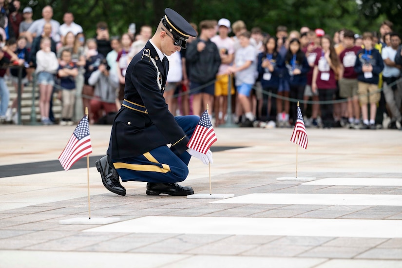 A man in a formal military uniform kneels while placing a small American flag into a flag holder on the ground.