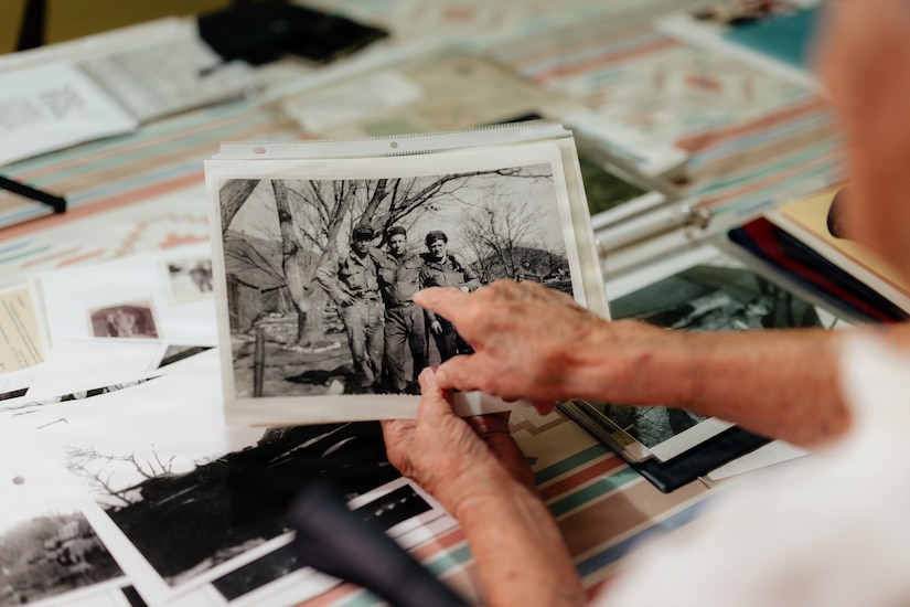 An older man points to a photo in his other hand of three men in military uniforms posing for a photo outside.
