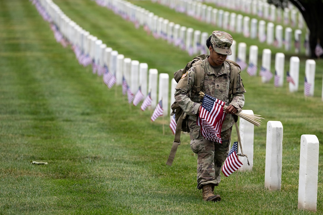 A soldier carries small U.S. flags in a cemetery, where a identical flags adorns each white headstone.