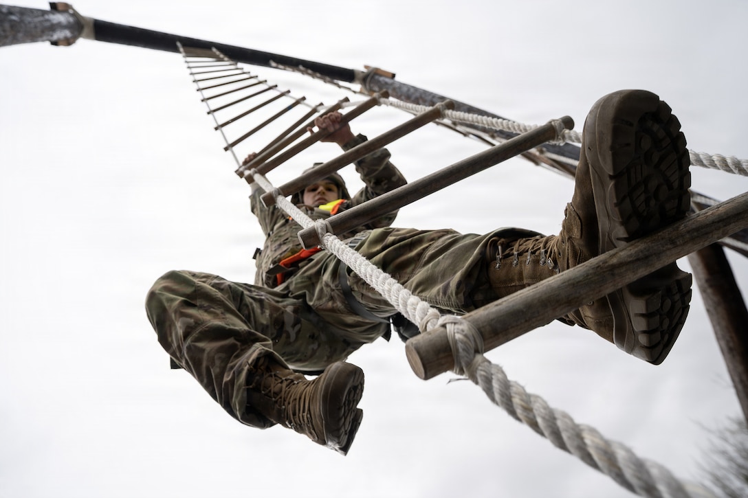 A soldier, viewed from below, climes a rope ladder with wooden rungs.