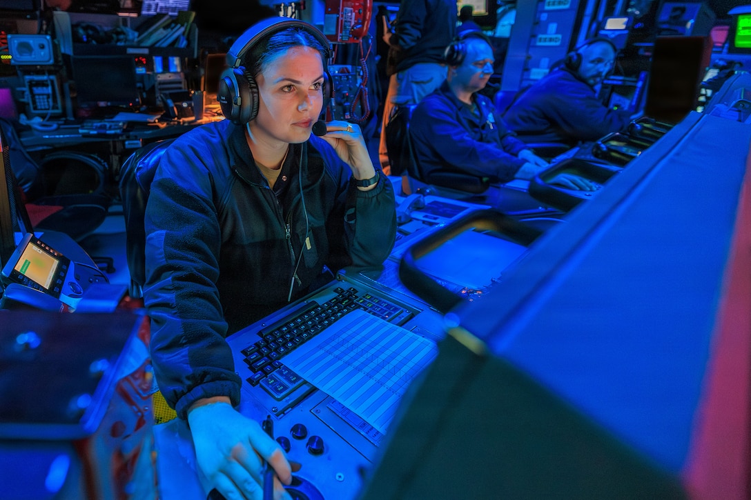 Sailors wearing headphones sit at computer terminals in a blue-lit room.