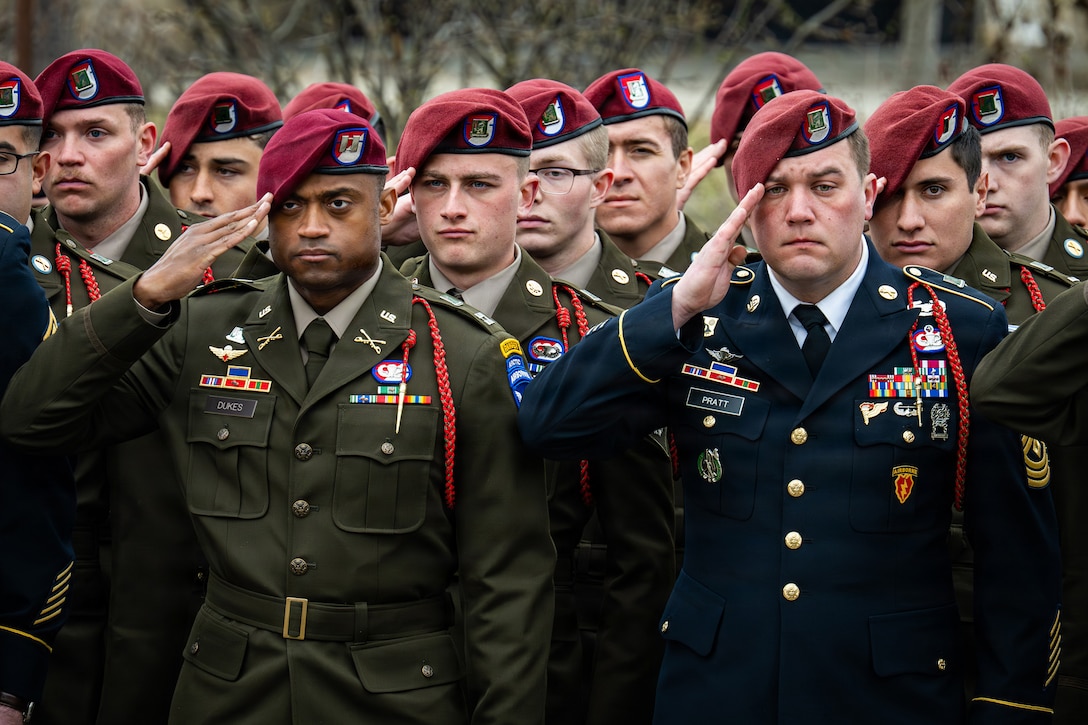 Soldiers in dress uniforms and burgundy berets stand and salute in rows outdoors.