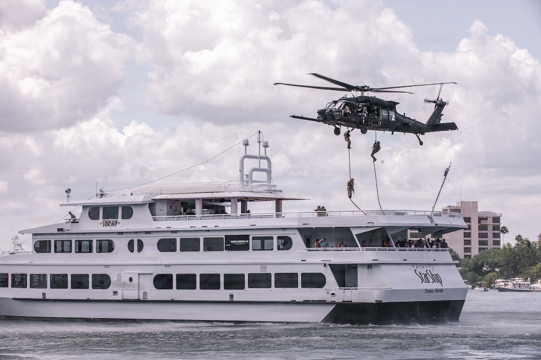 Service members descend on ropes from an Army helicopter hovering over a white boat near a shoreline.