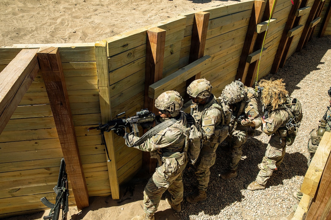 Four soldiers, seen from above, carry weapons while walking in a line along the wood wall of a trench.