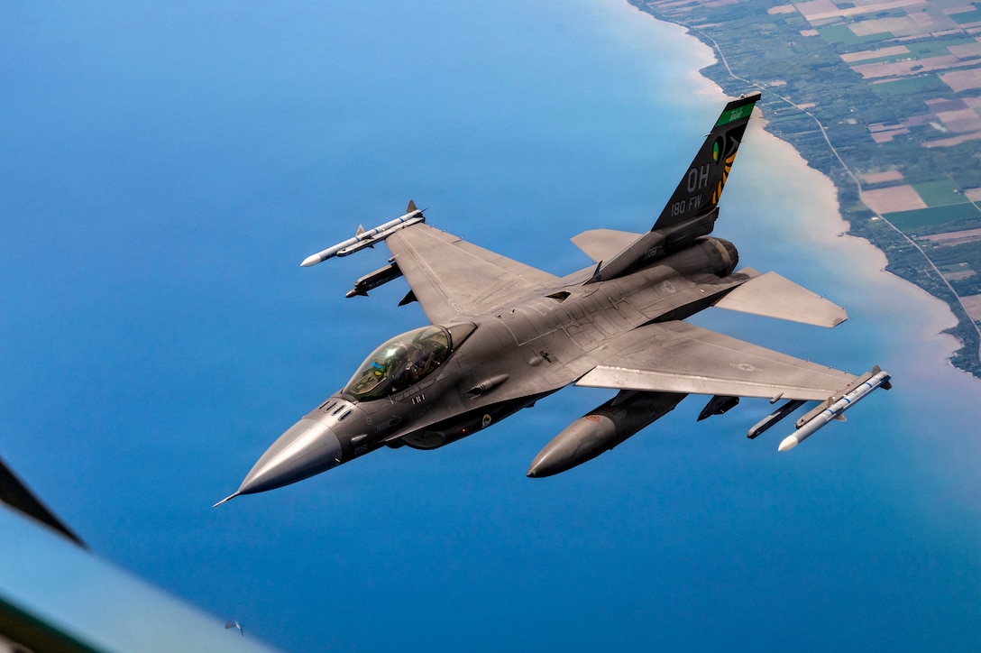 An Air Force fighter jet flies over blue water off a green shoreline.