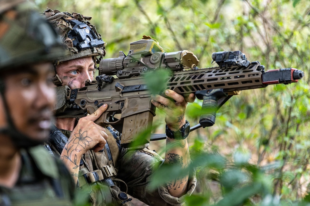 A soldier looks through the scope of a weapon while standing net to another service member in a forested area.