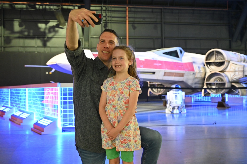 A man kneels next to a young girl with his cell phone held in front of them taking a selfie of them in front of one of the Iconic Starfighters on display at the National Museum of the U.S. Air Force.