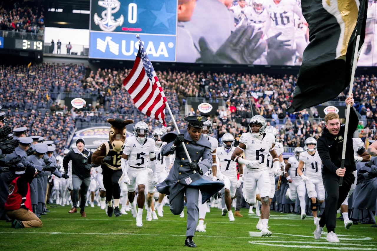 Two people, one in a military dress uniform, run on a field while holding flags in front of a team of football players in uniform, with a large screen and spectators in the stadium in the background.
