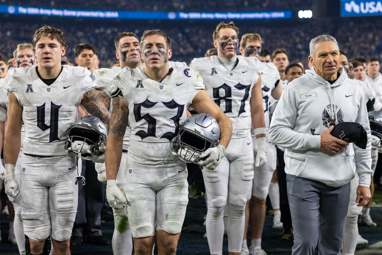 Football players in uniform and holding their helmets stand on a field with a person in a hooded sweatshirt and sing.