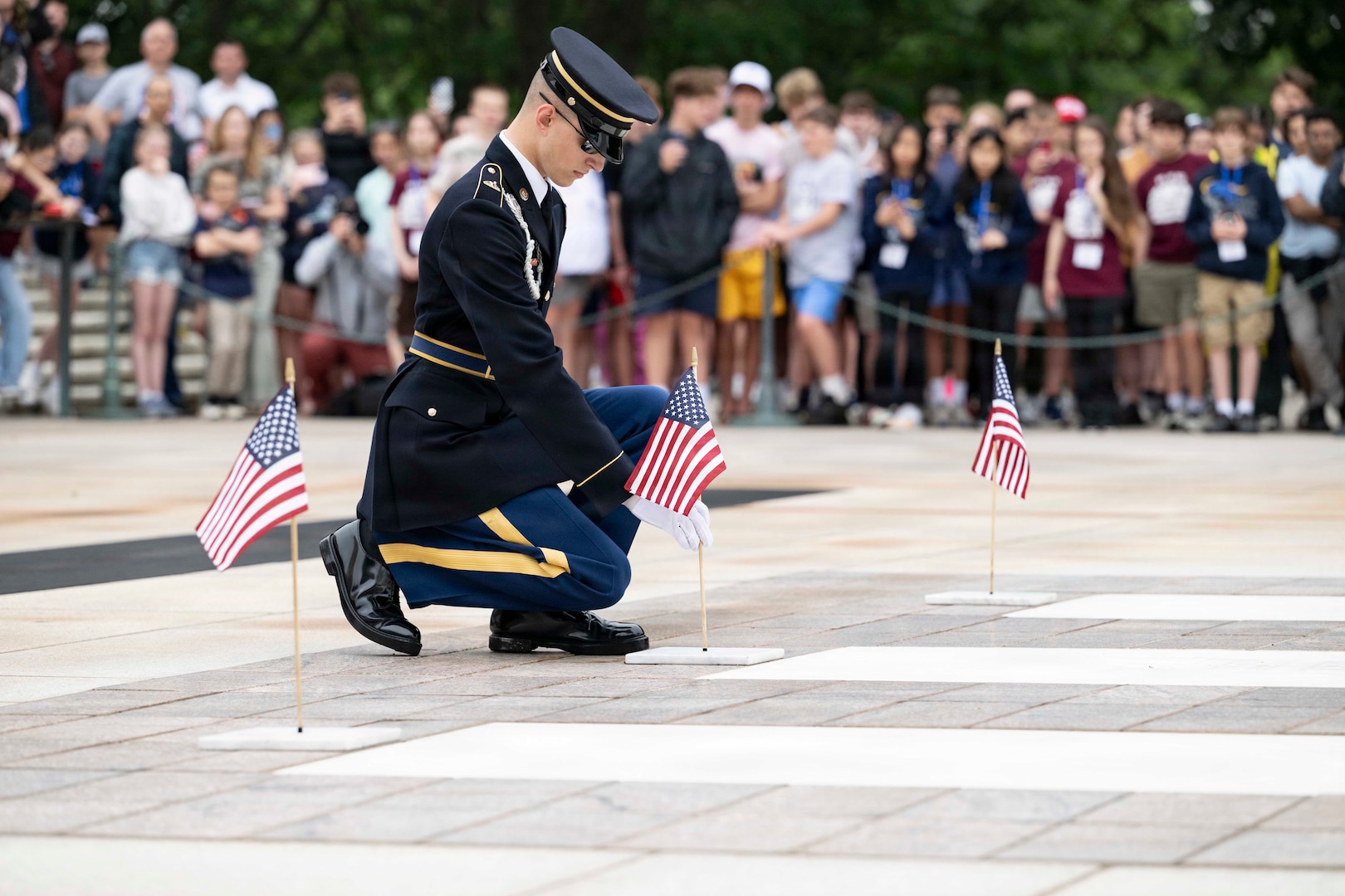 A man in a formal military uniform kneels while placing a small American flag into a flag holder on the ground.