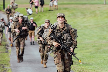 Cadets in military uniforms and carrying rifles march on a paved path during daytime, with other people in civilian attire also walking or standing in the background.
