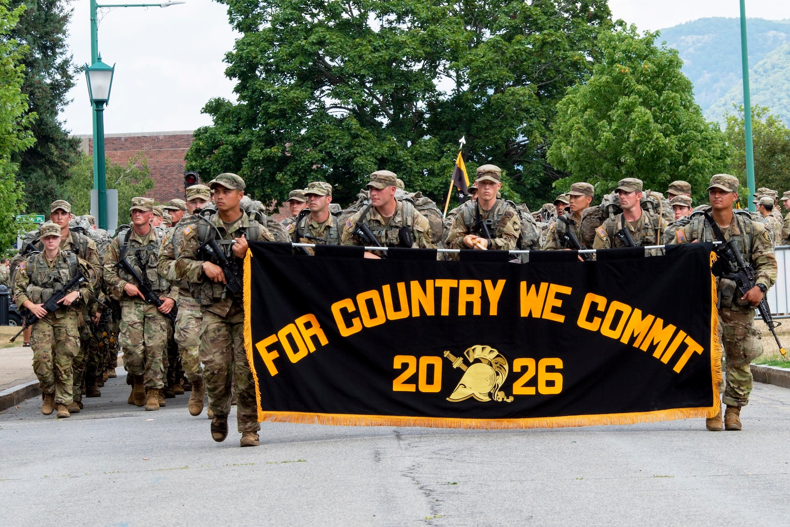 Soldiers wearing camouflage uniforms and carrying rifles march in formation on a road during daytime. The soldiers at the front hold a banner reading "For Country We Commit" and "2026."