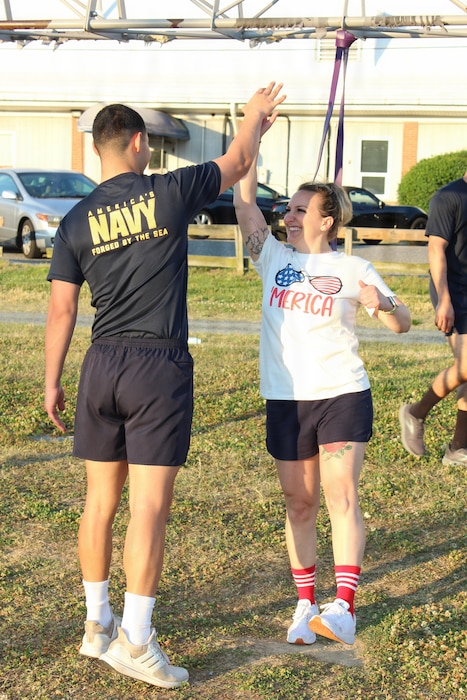 Assistant Command Fitness Leader Hospital Corpsman 3rd Class Lucas Arteaga, left, high-fives Command Fitness Leader Chief Hospital Corpsman Amanda Ellis at the Naval Air Station Patuxent River Drill Hall Field during the command’s monthly physical training event.

The two fitness leaders helped coordinate and drive motivation as Navy Medicine Readiness and Training Command (NMRTC) Patuxent River personnel took on a modified version of the 'Murph' challenge to foster unit cohesion and build camaraderie ahead of the Memorial Day weekend. Adding to the patriotic spirit of the morning, personnel wore either traditional Navy PT gear or red, white, and blue coordinated attire. 

The workout honors Navy SEAL Lt. Michael Murphy, who was killed in action in Afghanistan in 2005 and posthumously awarded the Medal of Honor. The traditional routine consists of a 1-mile run, 100 pull-ups, 200 push-ups, 300 squats, and a final 1-mile run.