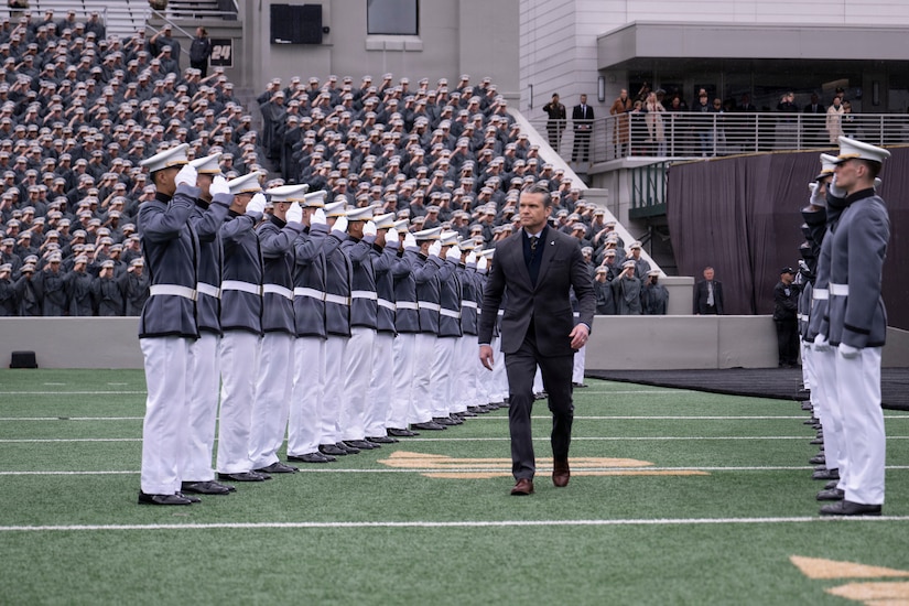 A man in business attire walks between two rows of more than a dozen saluting people in formal military uniforms, outside in a stadium; behind them, people in similar attire stand in the audience.
