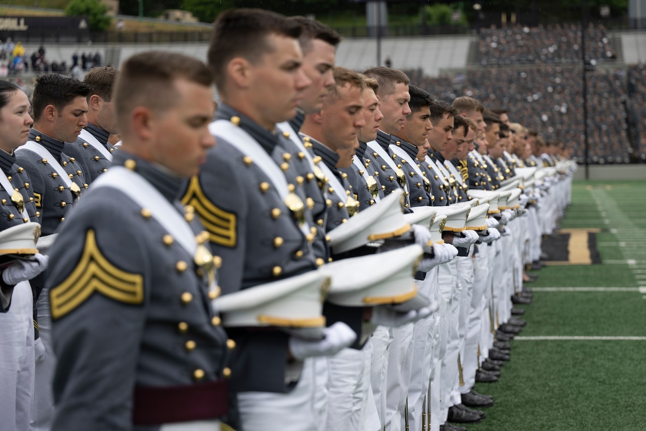 More than a dozen people in formal military uniforms bow their heads and holds their caps while standing in rows.