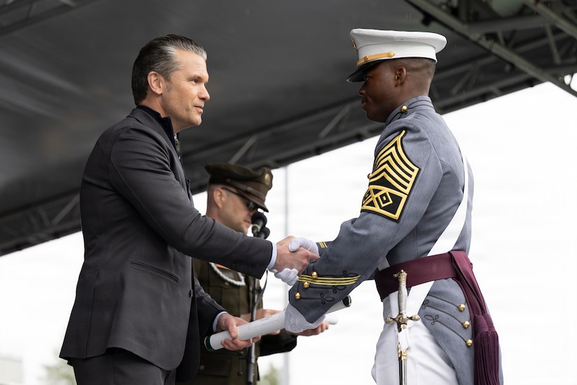 A man in business attire shakes hands with a man in a formal military uniform outside under a tent.