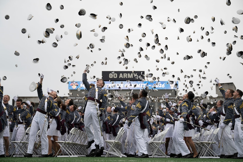 Hundreds of people in formal military uniforms throw hats into the air, outside in a stadium.