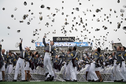 Hundreds of people in formal military uniforms throw hats into the air, outside in a stadium.