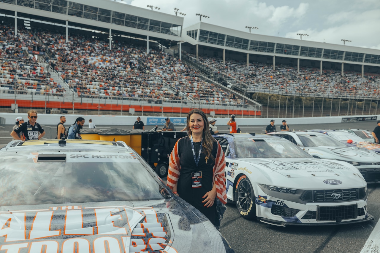 A woman in casual attire poses next to a race car on a track. Hundreds of people are in the stands behind her.