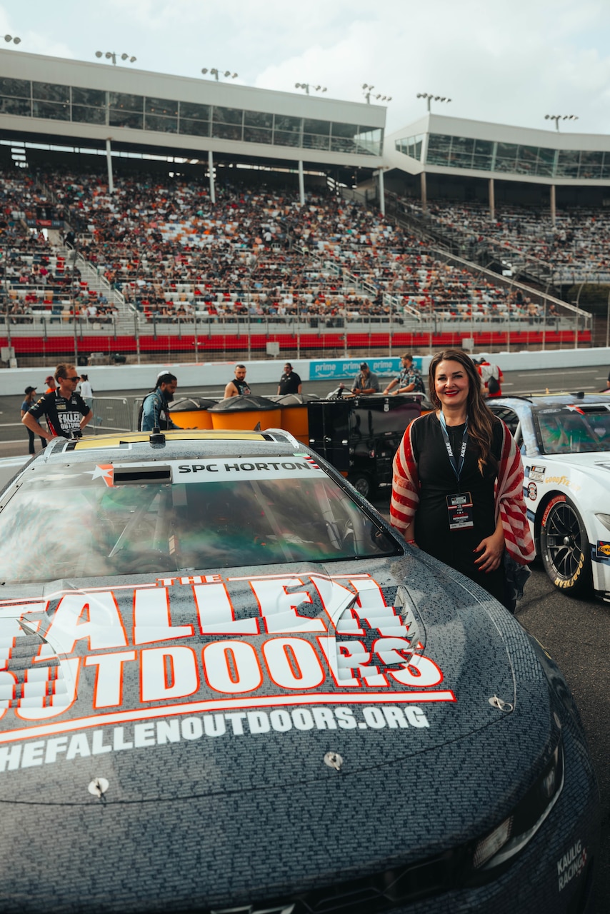 A woman in casual attire poses next to a race car on a track. Hundreds of people are in the stands behind her.