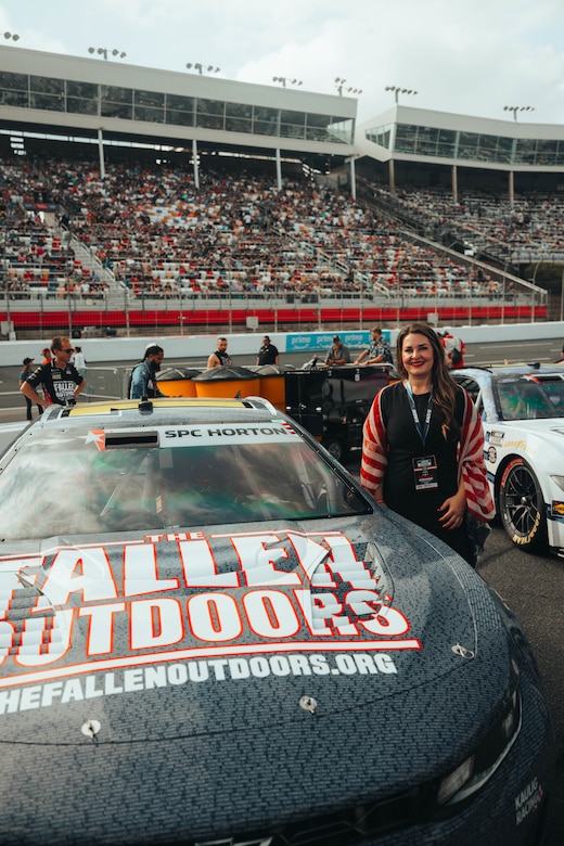 A woman in casual attire poses next to a race car on a track. Hundreds of people are in the stands behind her.
