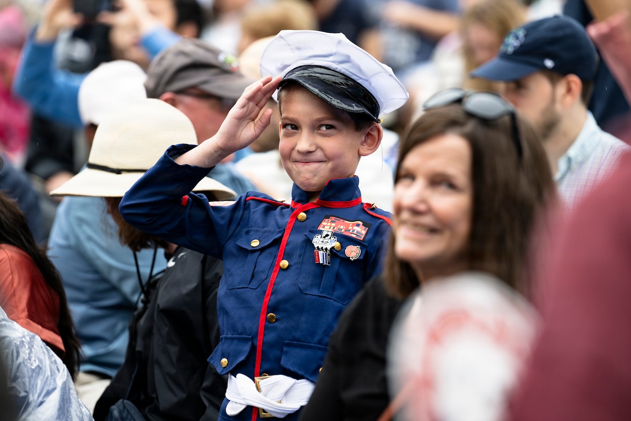 A little boy in a military uniform costume smiles and salutes in a crowd.