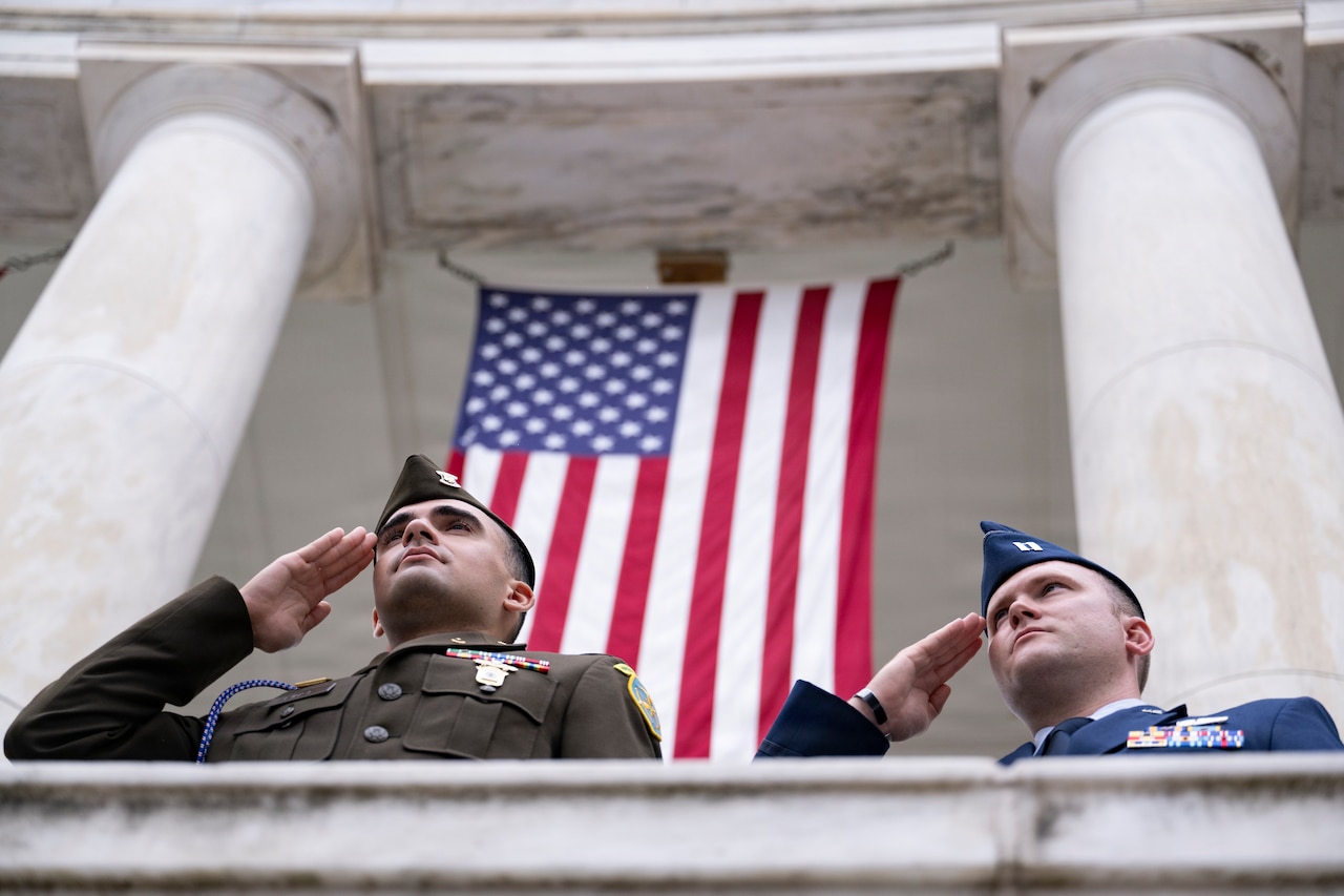 Two men in formal military uniforms render salutes outside a stone building, with an American flag in the background.
