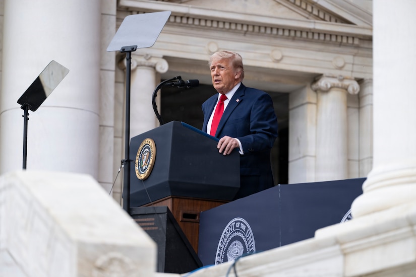 A man in business attire stands behind a lectern and speaks into a microphone outside a stone building.
