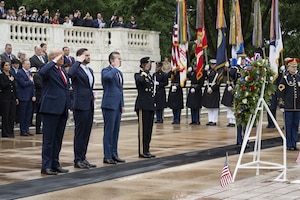 Three people in business attire and one in a formal military uniform stand and salute a wreath; behind them, people in similar attire have their hands over their hearts.