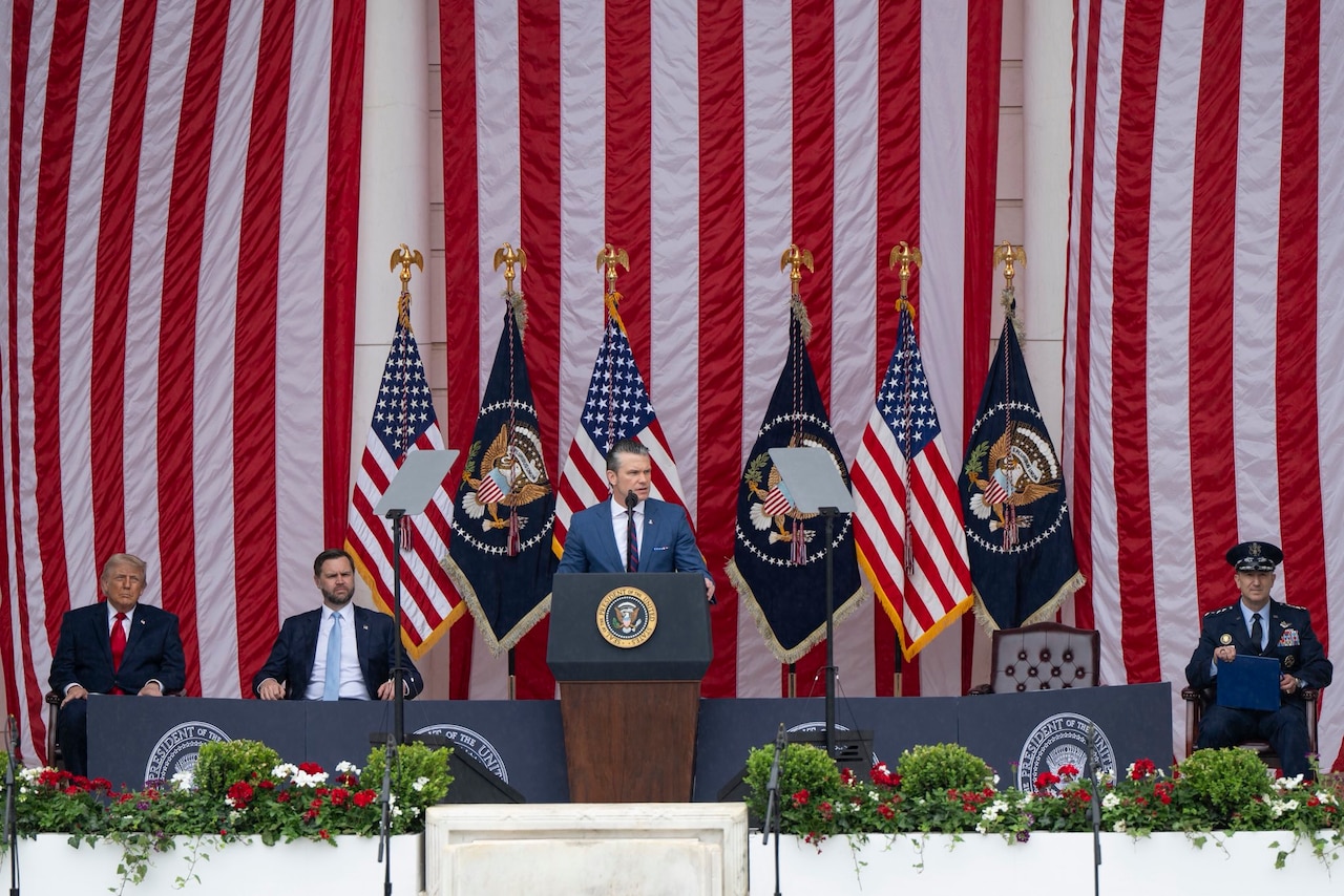 A man in business attire stands at a lectern and speaks into a microphone. Behind him are various flags and three seated men in similar attire.