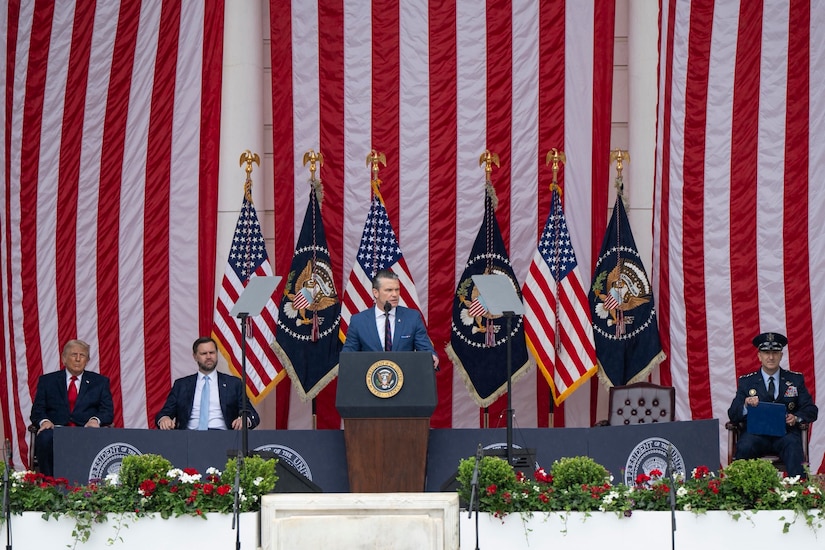 A man in business attire stands at a lectern and speaks into a microphone. Behind him are various flags and three seated men in similar attire.