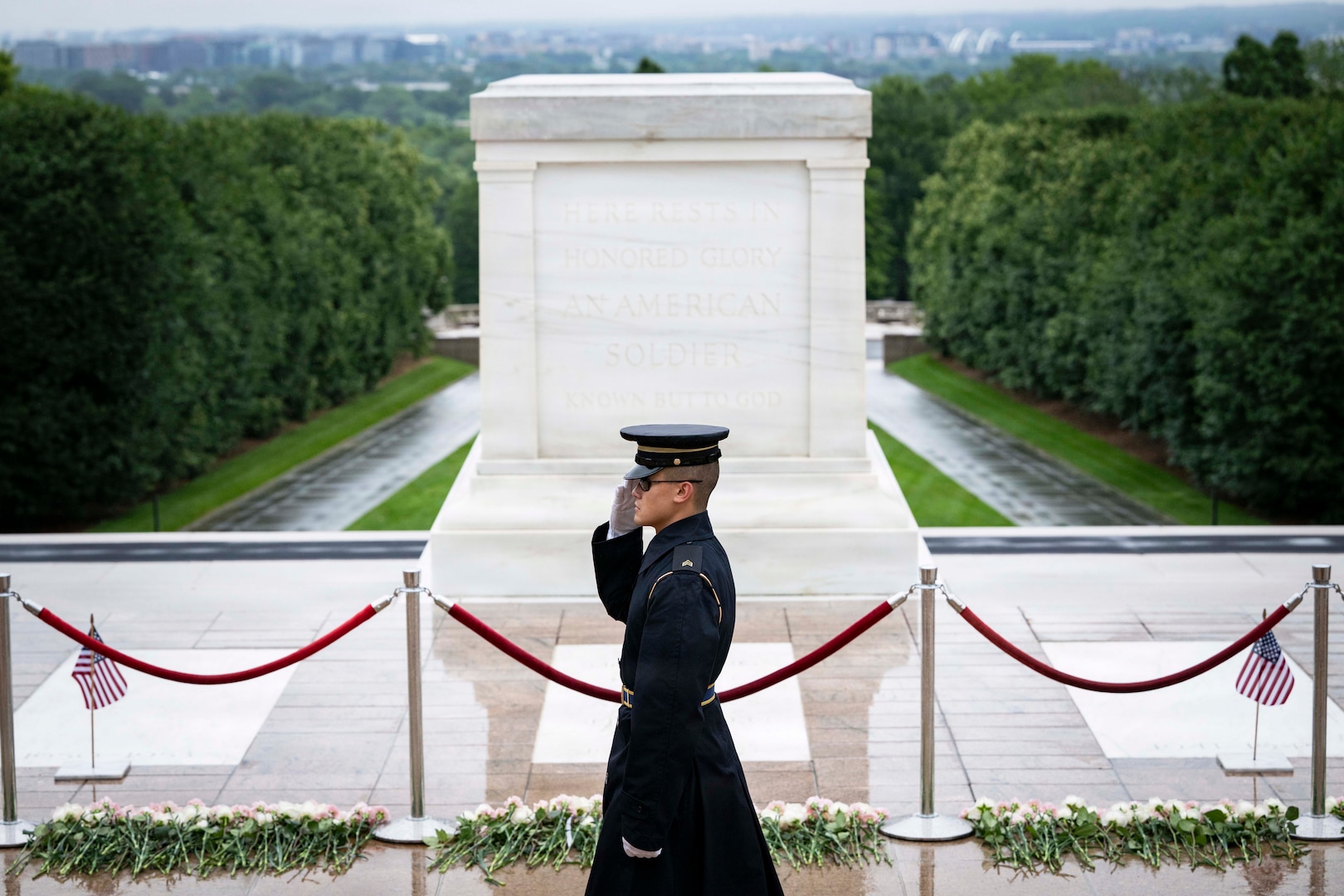 A soldier in a military dress uniform salutes as he walks in front of a large tomb and three gravestones during daytime, with trees in the background.