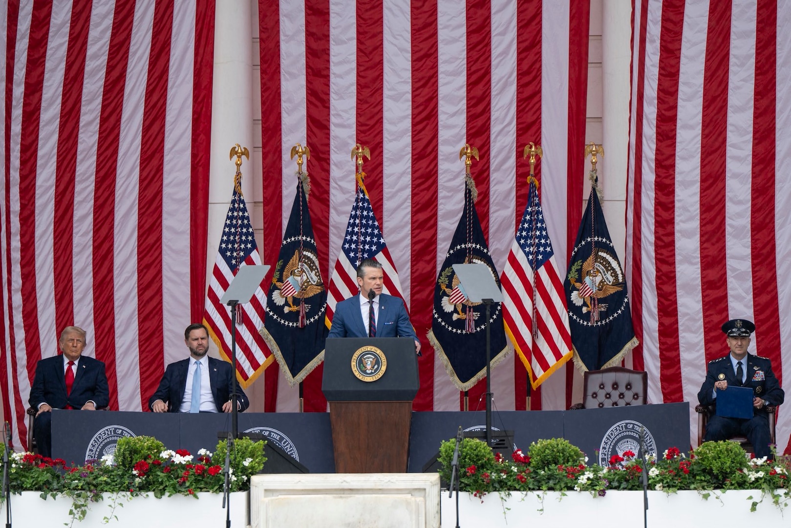 A man in business attire stands at a lectern and speaks into a microphone. Behind him are various flags and three seated men in similar attire.
