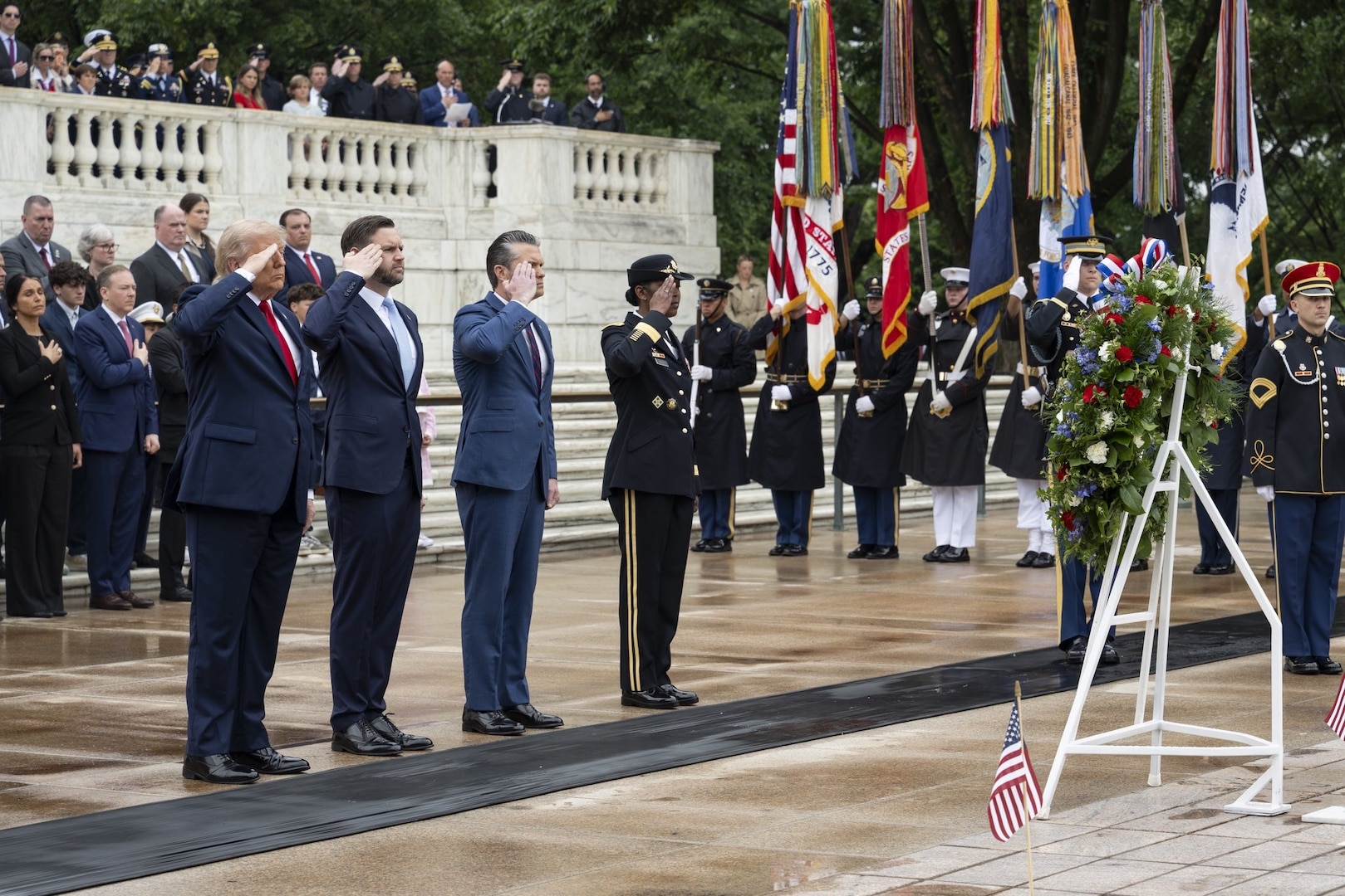 Three people in business attire and one in a formal military uniform stand and salute a wreath; behind them, people in similar attire have their hands over their hearts.