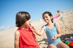 Two children sit on a beach while one applies sunscreen on the other's face.