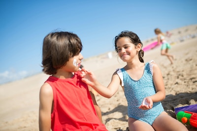 Two children sit on a beach while one applies sunscreen on the other's face.