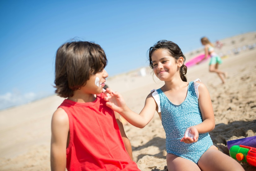 Two children sit on a beach while one applies sunscreen on the other's face.