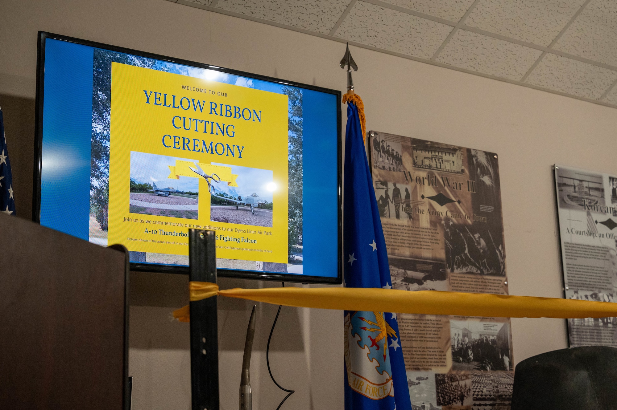 A ceremonial ribbon is positioned in front of a television welcoming guests to a yellow ribbon-cutting ceremony inside the Dyess Memorial Museum at Dyess Air Force Base, Texas, May 21, 2026. The dedication marked the completion of a three-year effort by the 7th Civil Engineer Squadron and the Dyess Memorial Museum to preserve the heritage of the newly installed A-10 Thunderbolt II and F-16 Fighting Falcon static displays. (U.S. Air Force photo by Airman 1st Class William Neal)