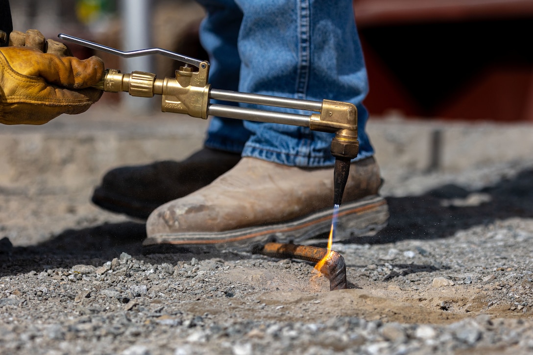A person wearing work boots and a leather glove uses a torch to heat the end of a metal rod sticking out of the gravel-covered ground.
