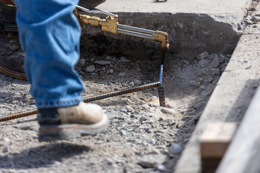 A construction worker in jeans and boots uses a torch to heat a piece of rebar inside a concrete trench.