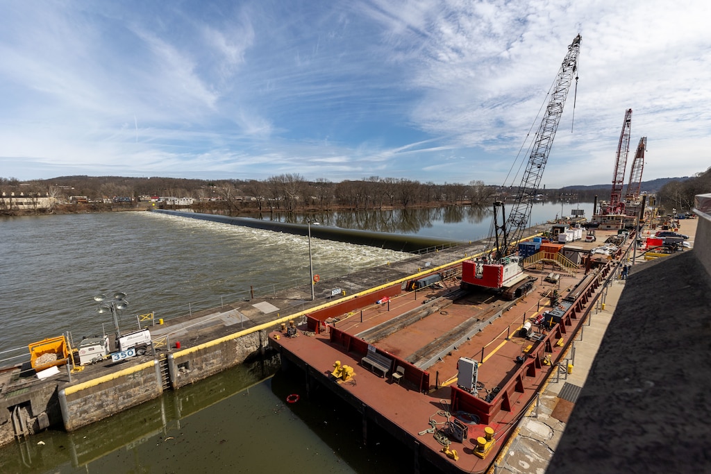 A wide, high-angle view of a river lock and dam with a large crane on a barge in the foreground.