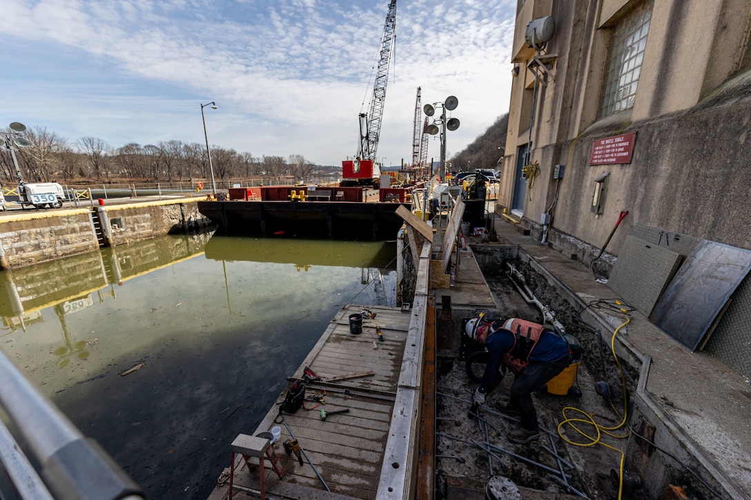 A construction worker in a trench next to a canal lock, with a barge and crane in the water.