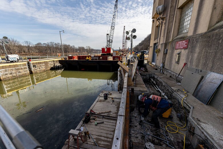 A construction worker in a trench next to a canal lock, with a barge and crane in the water.