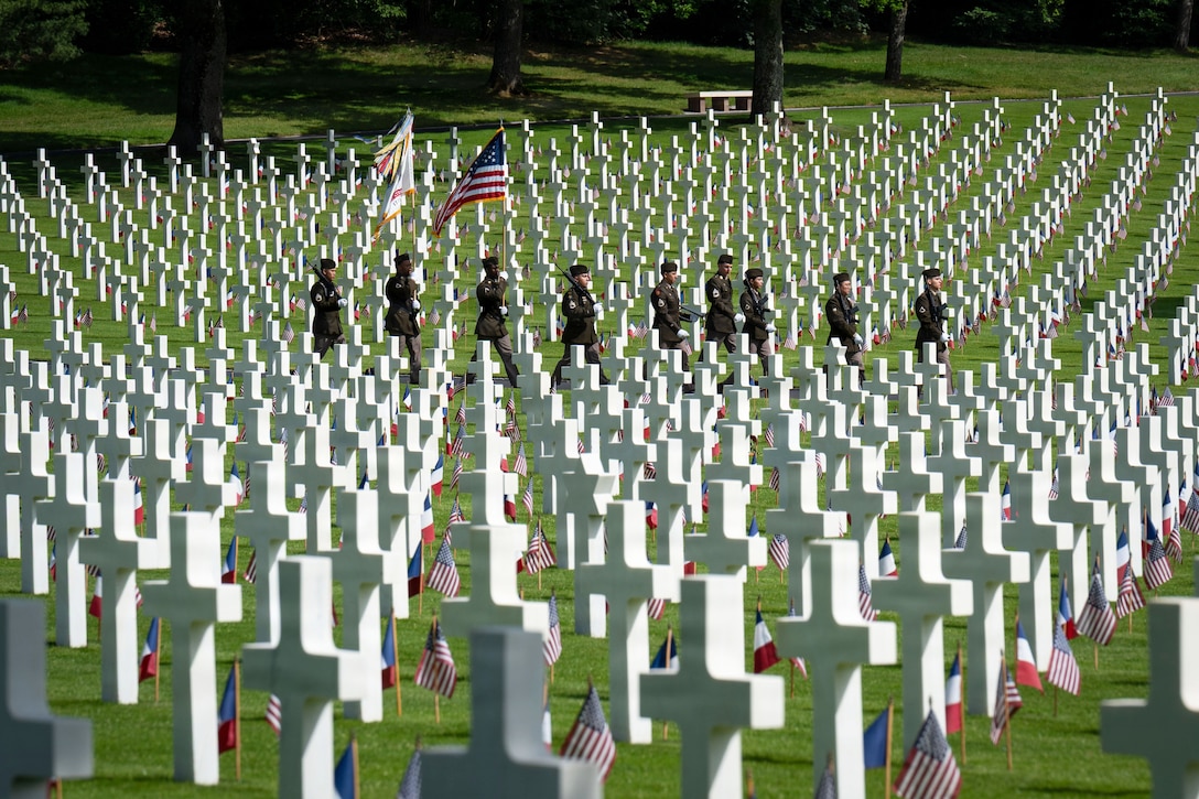Soldiers in ceremonial dress march in formation through a cemetery with dozens of cross tombstones and American flags.