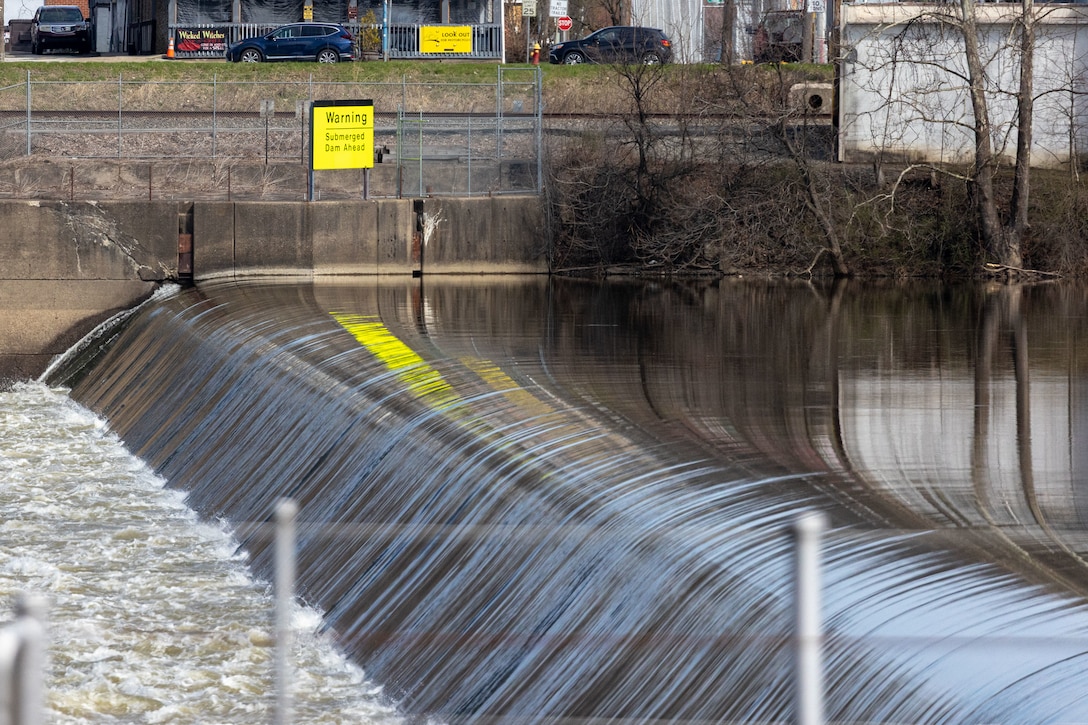 A view of a dam on a river, with water flowing smoothly over the edge. A yellow warning sign is visible on the concrete barrier of the dam.